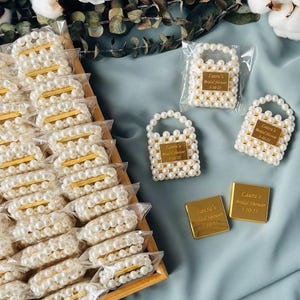 Op de afbeelding: Een verzameling met parel-geparelde mini-handtassen en goudkleurige rechthoekige labels, sommige verpakt in doorzichtig plastic. De labels en handtassen hebben de tekst "Laura's Bridal Shower 5-10-25".