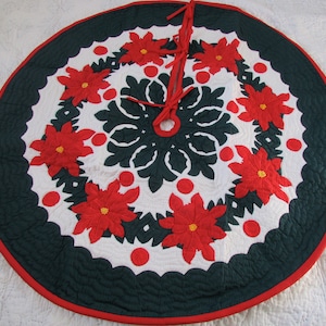 May include: A green and white quilted Christmas tree skirt with a red trim. The skirt features a snowflake design in the center and poinsettia flowers around the edge.