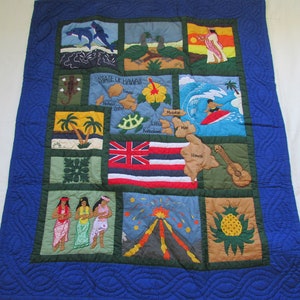 May include: A blue quilted wall hanging with a patchwork design featuring various Hawaiian themes, including a map of the islands, flowers, and surfers. The text "STATE OF HAWAII" is embroidered in the center.