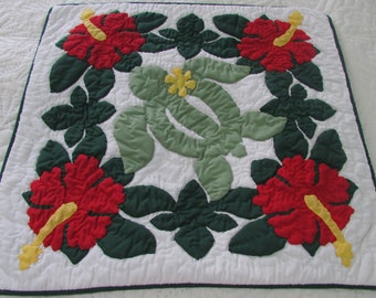 May include: A white quilted pillow with a green, red, and yellow floral design. The design features a turtle in the center surrounded by hibiscus flowers and leaves.