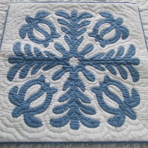 May include: Square white and blue quilt with a central blue floral design and four blue turtle motifs. The quilt has a blue border and is likely handmade. The quilting pattern is visible in the white background.