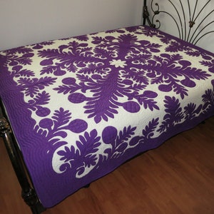 May include: A purple and white Hawaiian quilt with a floral design. The quilt is on a bed with a black metal frame.