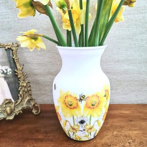 May include: A white vase with a floral design featuring yellow sunflowers and bees. The vase holds a bouquet of yellow daffodils with green stems and leaves. A gold framed picture is on the left.
