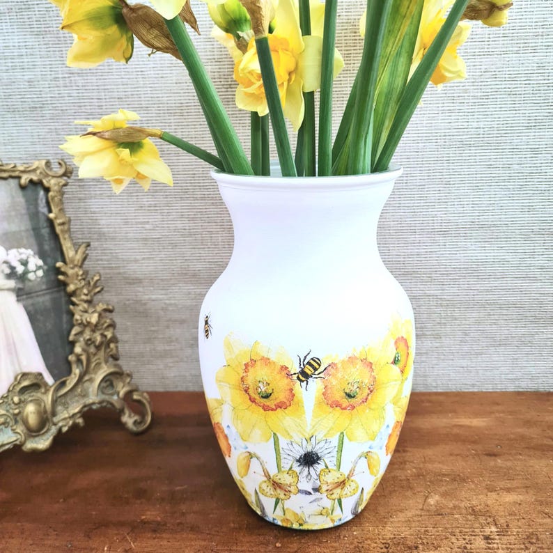 May include: A white vase with a floral design featuring yellow sunflowers and bees. The vase holds a bouquet of yellow daffodils with green stems and leaves. A gold framed picture is on the left.