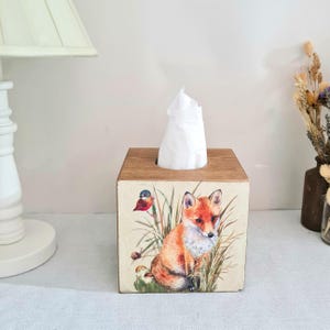 May include: Wooden tissue box with a fox and bird illustration. The box is square with a light brown wood finish on the top and sides. The illustration features a red fox, a small bird, and tall grass. White tissues are visible.