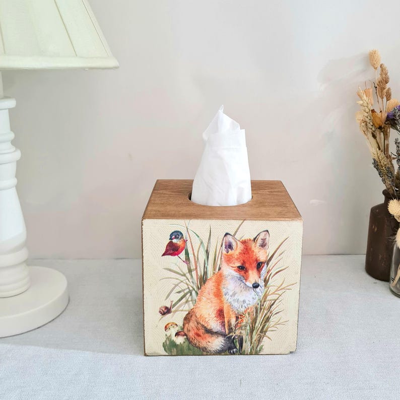 May include: Wooden tissue box with a fox and bird illustration. The box is square with a light brown wood finish on the top and sides. The illustration features a red fox, a small bird, and tall grass. White tissues are visible.