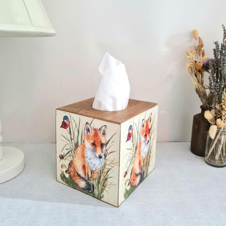 May include: A wooden tissue box featuring a fox design. The box has a light brown wooden top and sides decorated with illustrations of foxes, birds, and foliage. White tissues are visible at the top.