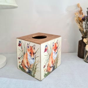 May include: Square wooden tissue box featuring a fox and bird design. The box has a light-colored background with illustrations of foxes, birds, and foliage. The top of the box is a natural wood color, and the tissue opening is a circular hole.