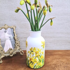 May include: A white vase with yellow daffodil floral design, holding green-stemmed daffodils with yellow buds. A gold-framed picture of a woman in a white dress is to the left. The vase is on a wooden surface.