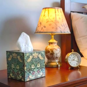 May include: A patterned tissue box, a white tissue, a gold lamp with a floral lampshade, and a small gold alarm clock are on a wooden bedside table. The lamp and clock add a touch of elegance. The tissue box has a green and gold floral design.