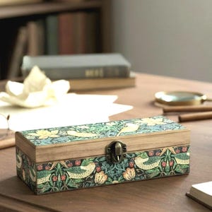 May include: A rectangular wooden box with a hinged lid featuring a metal latch. The exterior is decorated with a detailed botanical pattern in shades of green, cream, and red, depicting stylised birds, flowers, and foliage across the surface.