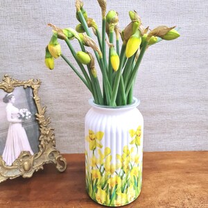 May include: A white vase with a yellow daffodil design holds a bouquet of daffodils with green stems and yellow buds. The vase has vertical ridges and sits on a wooden surface. A framed vintage photograph is to the left.