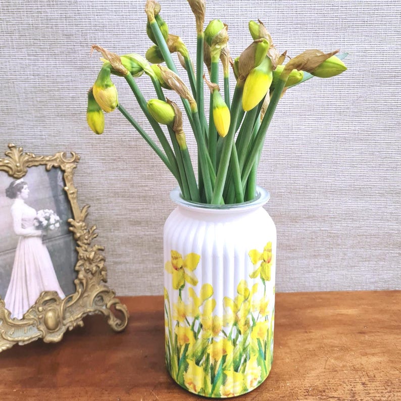 May include: A white vase with a yellow daffodil design holds a bouquet of daffodils with green stems and yellow buds. The vase has vertical ridges and sits on a wooden surface. A framed vintage photograph is to the left.