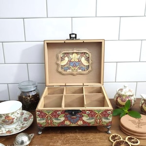 May include: A wooden tea box with six compartments, decorated with a floral and bird pattern on the front and lid. The box has small metal feet and a black clasp. A teacup, saucer, and tea infuser are visible, alongside a jar of tea.