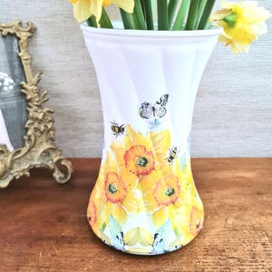 May include: White vase with a flared shape, decorated with yellow daffodils, butterflies, and bees. The vase has a striped pattern and is filled with yellow daffodils. A decorative item for the home.