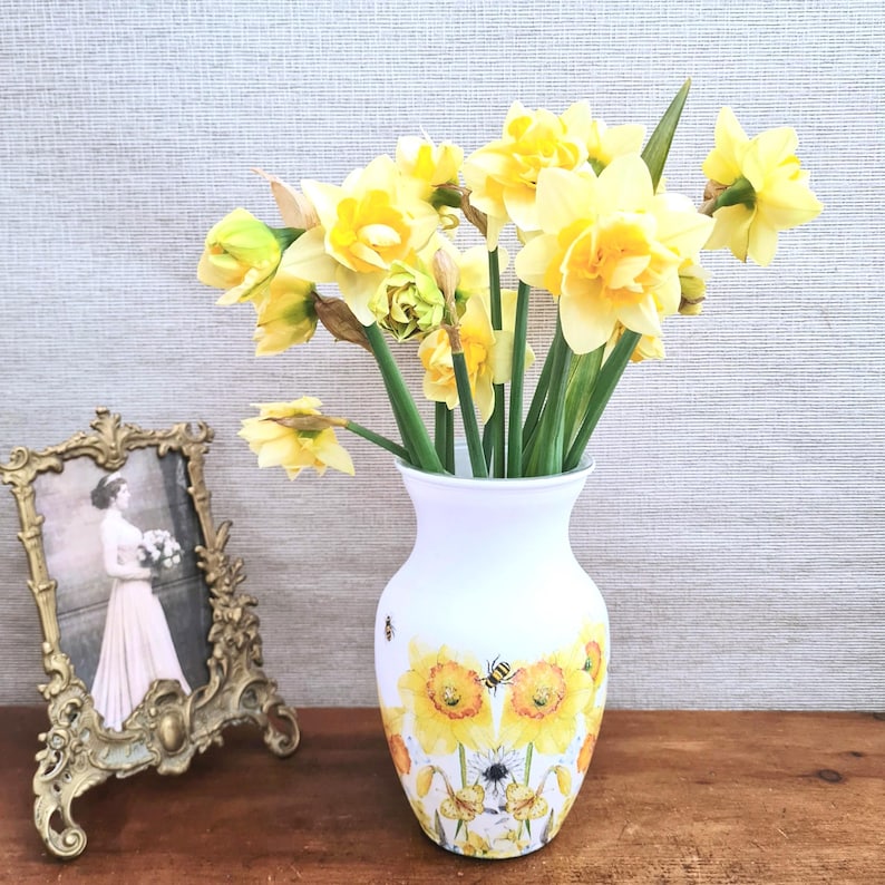 Spring daffodils tulip shaped glass vase, A pretty glass vase decoupaged with vibrant yellow daffodils with butterflies and bees amongst the flowers adding a touch of charm. against a hand painted in white chalk paint background giving a lovely rustic country feel with natural twine tied around the neck of the vase height 20 cm width 14 cm sealed to protect