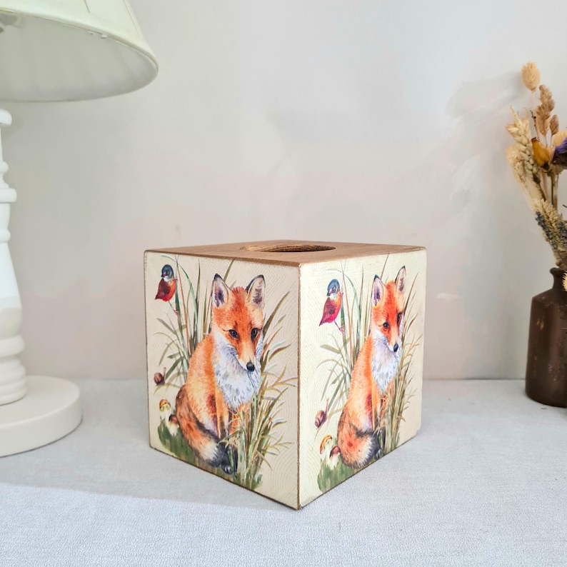 May include: A square wooden tissue box featuring a watercolor design of a fox in a grassy field with a bird. The box has a natural wood top and is painted with orange, brown, and green hues. The box is a decorative item.