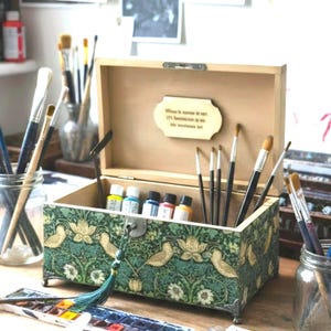 May include: A rectangular wooden storage box with a hinged lid, featuring a green and gold floral pattern with bird motifs. The interior lid displays a small plaque with text. The box contains several paintbrushes and small paint bottles.
