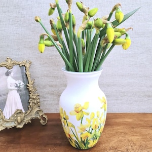 May include: A white vase with a yellow daffodil design holds a bouquet of green-stemmed daffodils with yellow buds. A framed black and white photo with a gold frame sits to the left of the vase on a wooden surface.