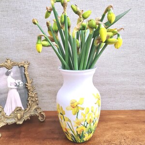 May include: A white vase with a yellow daffodil design holds a bouquet of fresh daffodils. The vase is on a wooden surface, and a framed photograph is visible in the background. The daffodils have green stems and yellow blooms.