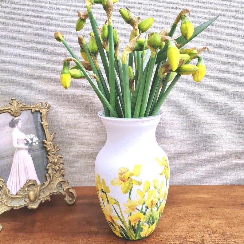 May include: A white vase with a yellow daffodil design holds a bouquet of fresh daffodils. The vase is on a wooden surface, and a framed photograph is visible in the background. The daffodils have green stems and yellow blooms.