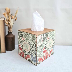 May include: A decorative tissue box with a wooden lid and sides adorned with a floral design in red, green, and blue. White tissues are visible. A stylish home accessory.