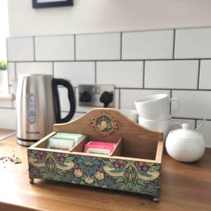 May include: Wooden tea bag storage box with a decorative floral and bird pattern. The box has three compartments, a decorative top, and small feet. Tea bags are visible inside. A kettle, cups, and sugar bowl are in the background.