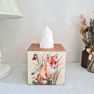 May include: Wooden tissue box featuring a painted fox, bird, and foliage design. The box is square with a natural wood top and a cream-colored background for the artwork. A stack of white tissues is visible.