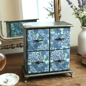 May include: A rectangular wooden chest with four drawers, featuring a blue and green floral pattern. The chest has a dark top surface with decorative metal corner accents, small metal feet, and four small, dark, round drawer pulls.