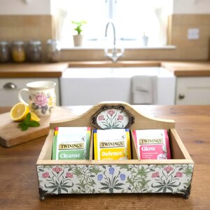 May include: A wooden tea caddy with three compartments, each holding a different flavour of Twinings tea. The caddy is decorated with floral patterns and has a decorative floral accent. A teacup, lemon, and mint sit nearby.