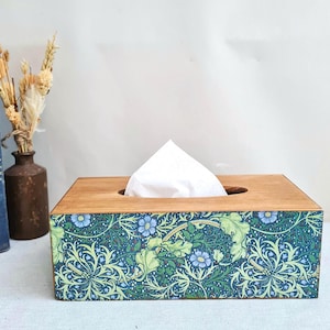 May include: Wooden tissue box featuring a floral design in blue and green hues. The box has a rectangular shape with a natural wood finish on the top and sides. White tissues are visible. A decorative home accessory.