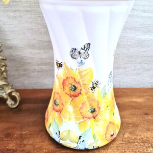 May include: White vase with a flared shape, decorated with yellow daffodils, butterflies, and bees. The floral design wraps around the vase, creating a spring-like aesthetic. The vase is set on a wooden surface.