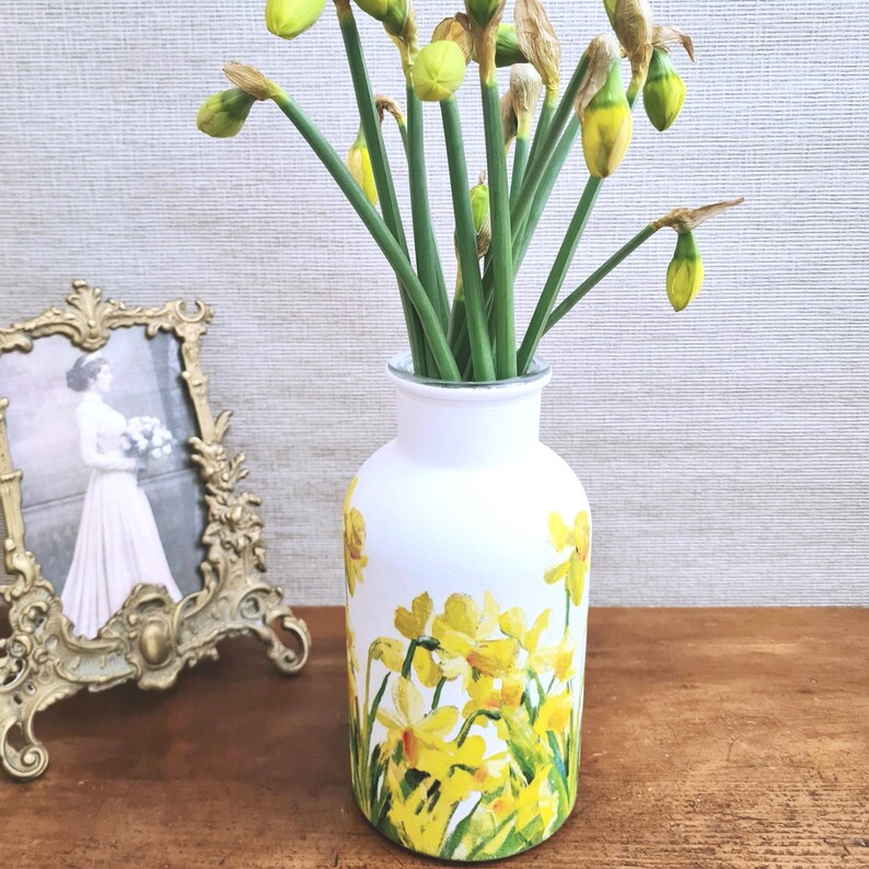 Spring Daffodils glass vase, A beautiful glass vase which has been decoupaged with vibrant yellow spring daffodils all around the vase creating a cheerful and uplifting display. set against a hand painted white chalk paint background giving it a rustic feel with natural twine tied around the vase. Height 20 cm width 11 cm
