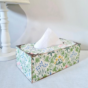 May include: A rectangular tissue box with a floral pattern of green leaves, blue and yellow flowers, and pink accents. The box has a decorative metal trim and a white tissue is pulled from the top opening. The box is on a white surface.