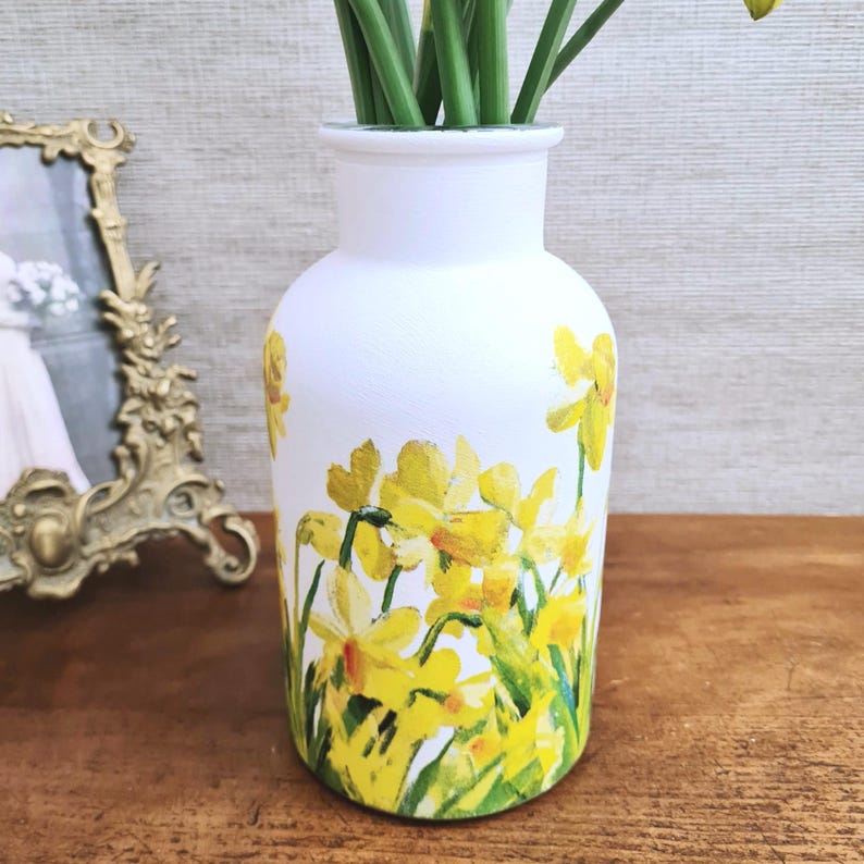 May include: White vase with a hand-painted design of yellow daffodils and green stems. The vase has a rounded shape and a narrow neck. The daffodils are clustered around the base of the vase. The vase is sitting on a wooden surface.