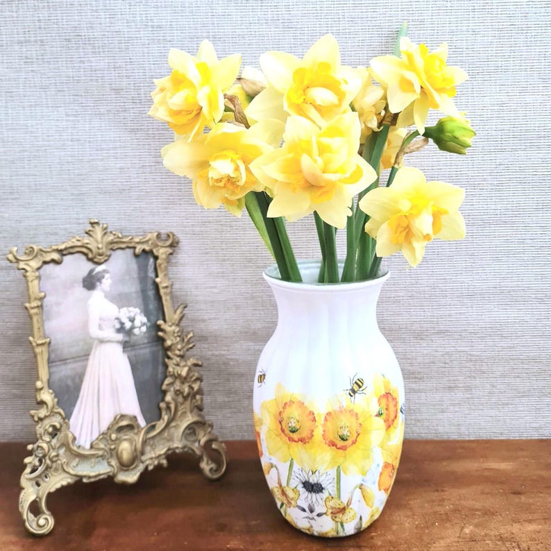 Spring daffodils tulip shaped glass vase, A pretty glass vase decoupaged with vibrant yellow daffodils with butterflies and bees amongst the flowers adding a touch of charm. against a hand painted in white chalk paint background giving a lovely rustic country feel with natural twine tied around the neck of the vase height 20 cm width 14 cm sealed to protect