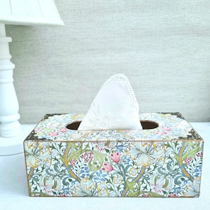 May include: A decorative tissue box featuring a floral design in green, blue, pink, and yellow. The box has metal corner detailing and a white tissue emerging from the top. The pattern is reminiscent of vintage decor, with a rectangular shape.