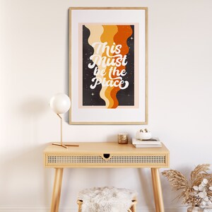 May include: A framed print with a retro design featuring a black background with orange and yellow wavy lines and white stars. The text "This Must Be The Place" is written in white cursive font.