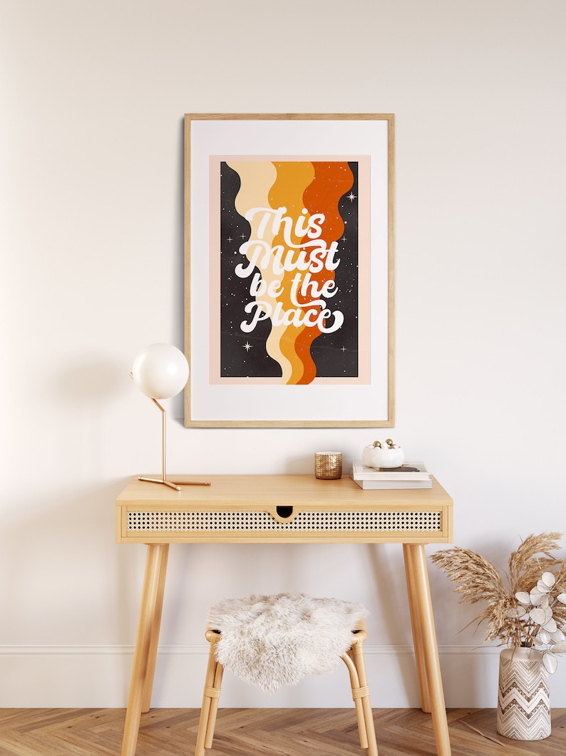 May include: A framed print with a retro design featuring a black background with orange and yellow wavy lines and white stars. The text "This Must Be The Place" is written in white cursive font.