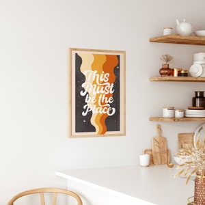 May include: A retro-style print with a black background and orange, yellow, and cream coloured wavy lines. The text "This Must Be The Place" is written in white cursive font.