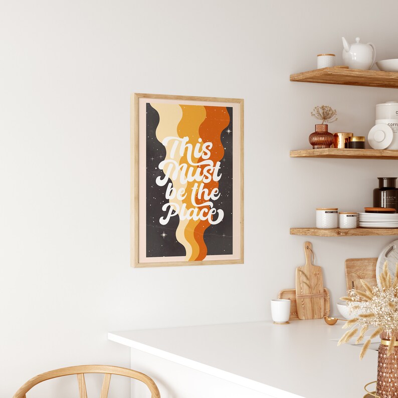 May include: A retro-style print with a black background and orange, yellow, and cream coloured wavy lines. The text "This Must Be The Place" is written in white cursive font.