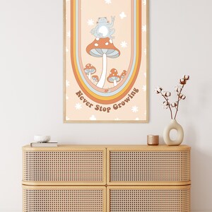 May include: A retro-style poster featuring a blue frog perched on a red mushroom. The poster has a rainbow arch and the text "Never Stop Growing" in brown lettering.