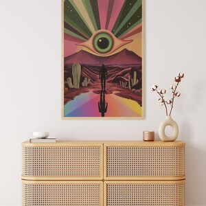 May include: A psychedelic art print featuring a large eye with a green iris and a brown pupil, surrounded by a rainbow of colours. The eye is set against a desert landscape with cacti and a lone figure walking towards it.