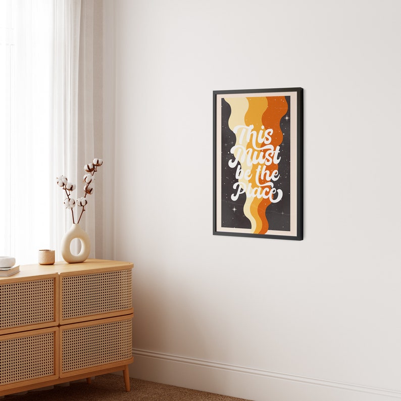 May include: A black framed poster with a retro design featuring a wavy orange, yellow, and brown abstract pattern on a black background with white stars. The text "This must be the place" is written in white cursive lettering.