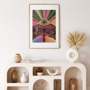 May include: A framed print of a psychedelic desert landscape with a large eye in the sky and a person walking towards it. The print is in a wooden frame and is hanging on a white wall.
