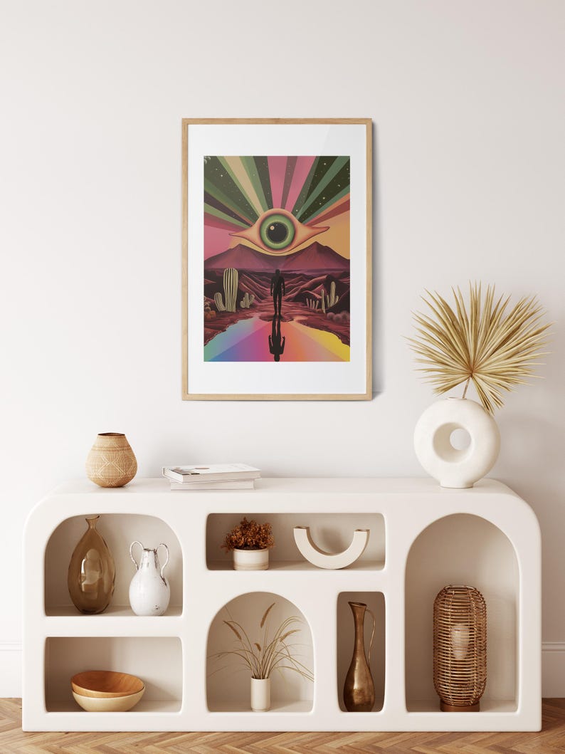 May include: A framed print of a psychedelic desert landscape with a large eye in the sky and a person walking towards it. The print is in a wooden frame and is hanging on a white wall.