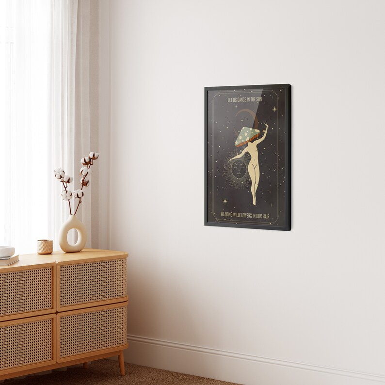 May include: A black framed print with a dark background and a woman dancing with a mushroom on her head. The text on the print reads "Let us dance in the sun, wearing wildflowers in our hair."