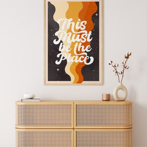 May include: A framed print with a retro design featuring a black background with orange, yellow, and brown wavy lines. The text "This Must Be The Place" is written in white cursive font.