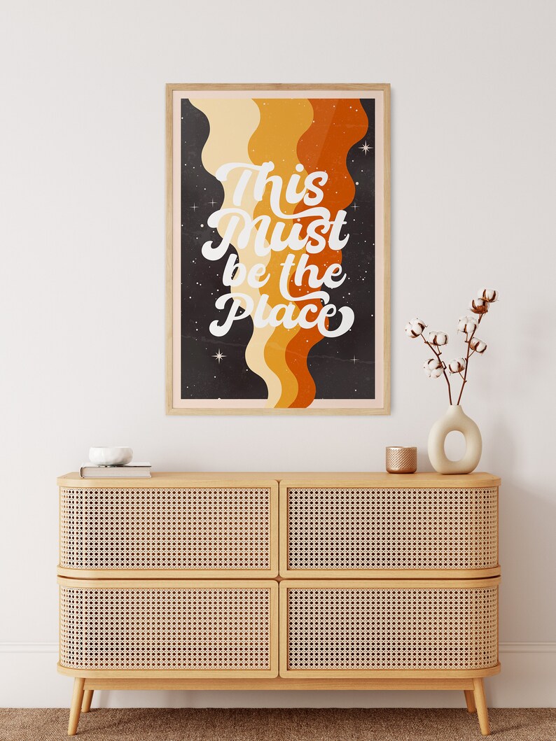 May include: A framed print with a retro design featuring a black background with orange, yellow, and brown wavy lines. The text "This Must Be The Place" is written in white cursive font.