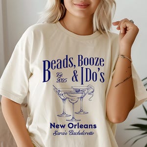 Puede incluir: Una camiseta blanca con un gráfico azul que dice "Beads, Booze & I Do's Est. 2025" con un dibujo de tres copas de martini. El texto "New Orleans Sara's Bachelorette" está debajo del gráfico.